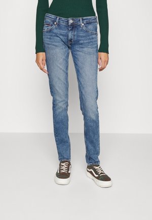 Persoon die strakke blauwe spijkerbroek, groene geribbelde trui en geruite sneakers draagt, staand tegen een effen lichte achtergrond.