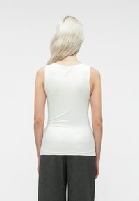 Personne avec de longs cheveux platine blonds tournés vers l'arrière, portant un haut sans manches blanc et un pantalon gris foncé sur un fond clair uni.