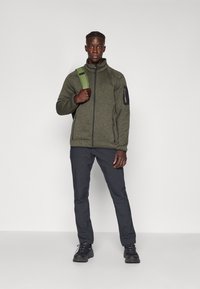 Grüne strukturierte Jacke mit Reißverschluss, schwarzen Akzenten, kombiniert mit dunklen Hosen und schwarzen Wanderschuhen. Rucksack mit grünem Gurt.