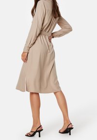Beżowa sukienka z długim rękawem, z paskiem w talii, z rozcięciem na dole i miękką teksturą. W zestawie z czarnymi sandałami na obcasie.