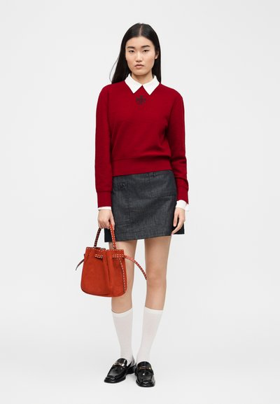 Jeune femme portant un pull rouge avec un col blanc, une jupe gris foncé, des chaussettes blanches montantes jusqu'aux genoux, des mocassins noirs, tenant un sac à main rouge.