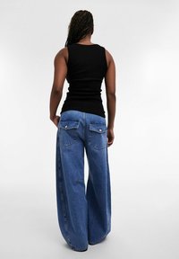 Personne aux cheveux tressés portant un débardeur noir et un jean bleu à jambes larges, debout dos à la caméra.