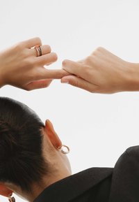 Dos manos entrelazadas, una adornada con anillos entrelazados en oro rosa y plata, la otra con piel suave; se ven pendientes.