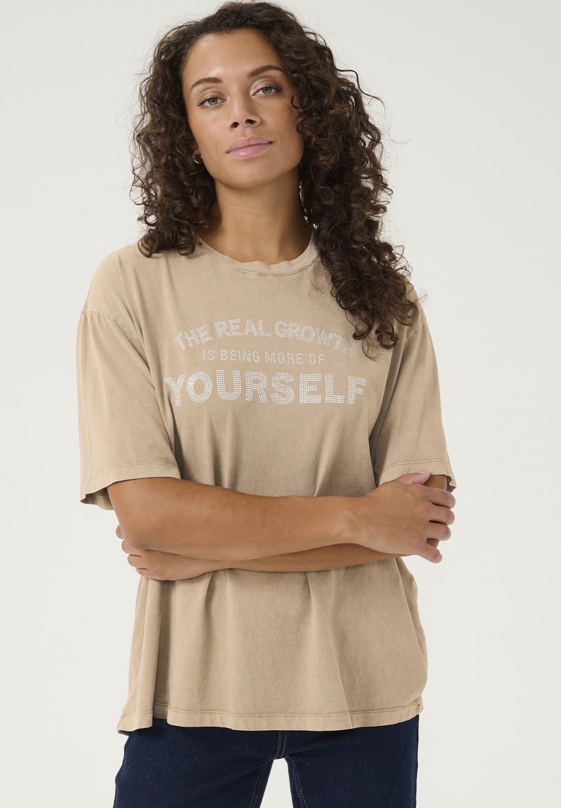 T-shirt en coton beige à manches courtes et col rond. Présente un imprimé texturé en blanc, indiquant "LA VRAIE CROISSANCE C'EST D'ÊTRE DAVANTAGE SOI-MÊME."