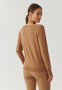Bruine gebreide trui met lange mouwen en een ronde halslijn, met een getailleerd ontwerp en een geribde zoom en manchetten. Draagt met beige broek.