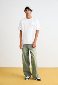 Bílá bavlněná trička s kapsou na hrudi, světle modrá kšiltovka a volné, vybledlé zelené džíny. Celý outfit doplňují černobílé tenisky.