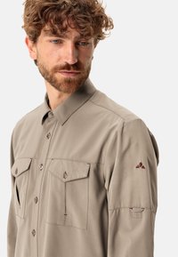 Camicia beige a maniche lunghe realizzata in tessuto morbido, con due tasche sul petto con chiusura a bottone e sottili dettagli di cucitura rossa.