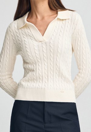 Femme portant un pull écru à torsades avec un col en V, associé à un pantalon noir taille haute sur un fond uni.