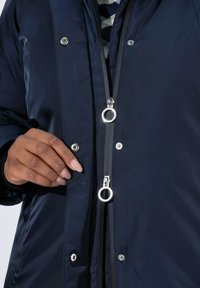 Chaqueta azul marino con una textura suave, que presenta un cierre con cremallera, herrajes plateados y botones a presión. Colocada a mano cerca de la cremallera.