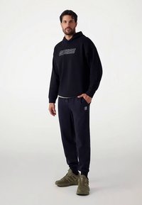 Czarna bluza z kapturem z logo "GUESS", zestawiona z granatowymi dresami z małym detalem w postaci logo. Dopełnieniem stroju są oliwkowe sneakersy.