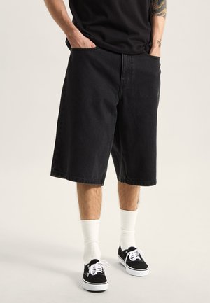 Shorts in denim nero dalla vestibilità morbida, lunghi fino al ginocchio, con tasche frontali. Abbinati a sneaker nere e calzini bianchi alla caviglia.