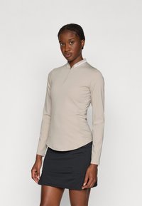 Maglia beige a maniche lunghe con colletto zip, abbinata a una gonna nera corta. Il tessuto sembra elastico e ha una superficie liscia.