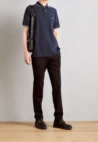 Granatowa koszulka polo z białą lamówką, w zestawieniu z czarnymi spodniami i czarnymi mokasynami. Modelka trzyma czarną torbę, stoi na drewnianej podłodze.