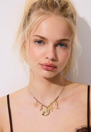 Giovane donna con capelli biondi indossa una collana d'oro con ciondoli a tema marino, tra cui una stella marina, una conchiglia e un pesce, sulle spalle scoperte con spalline nere.