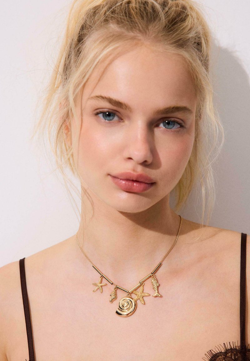 Jeune femme aux cheveux blonds portant un collier en or orné de charms sur le thème de la mer, incluant une étoile de mer, un coquillage et un poisson, sur des épaules nues avec des bretelles noires.
