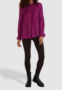 Lila pitkähihaisuus paita, jossa on pleateja, avokantinen kaula-aukko ja röyhelöhihojen päät, yhdistettynä mustiin leggingseihin ja nilkkureihin. Sujuva kankaan tekstuuri.