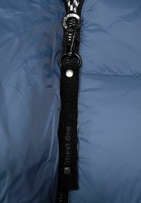 Blå puffjacka med en svart dragkedja med metallhalk, justerbar nackrem och silverdetaljer. Slät, texturerad tyg.