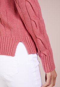 Pull rose tricoté avec un motif torsadé sur les manches, une texture côtelée et des fentes latérales, associé à un pantalon blanc.