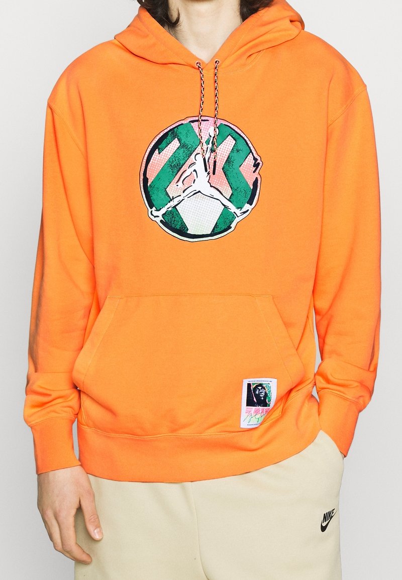 Personne portant un sweat à capuche orange vif avec un logo circulaire vert et rose ainsi qu'un écusson, associé à un pantalon de survêtement beige Nike.