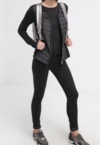 Svart pufferjacka med dragkedja över en svart topp, svarta tajta jeans och metallicfärgade sneakers; inkluderar en silverfärgad ryggsäck med justerbara remmar.