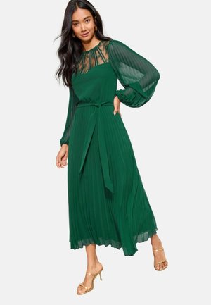 Grünes plissiertes Kleid mit durchsichtigen Ballonärmeln, Spitze vorne und gebundenem Taillengürtel. Kombiniert mit goldenen offenen High Heels. Glatter, fließender Stoff.