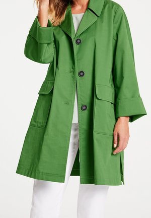 Femme portant un manteau vert vif longueur genou avec trois boutons noirs, grandes poches avant et poignets retroussés, par-dessus un haut rayé et un pantalon blanc.