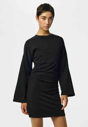 Jeune personne aux cheveux courts et foncés, portant une robe noire près du corps avec de longues manches larges, debout devant un fond clair uni.