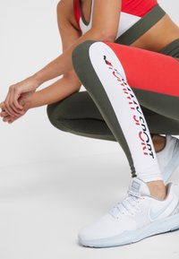 Leggings in Grün und Weiß mit einem roten Akzent, versehen mit einem "Sport"-Grafik. Kombiniert mit weißen Sportschuhen, die ein strukturiertes Design aufweisen.