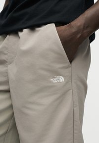 Δεξί χέρι στην τσέπη μπεζ παντελονιού με το λογότυπο The North Face, φορεμένο με μαύρο πουκάμισο σε απλό φόντο.