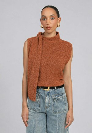 Femme portant un haut en tricot sans manches de couleur rouille avec un détail d'écharpe attaché, des boucles d'oreilles créoles dorées et un jean taille haute bleu clair avec des poches avant.