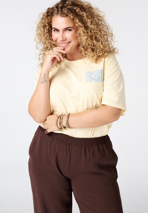 Femme aux cheveux blonds bouclés portant une chemise jaune et un pantalon marron, souriante, la main près de la bouche et plusieurs bracelets au poignet.