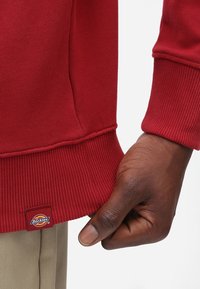 Felpa rossa con polsini e orlo a coste, presenta un piccolo etichetta con il logo Dickies sul lato. Tessuto morbido al tatto, primo piano di una mano che tiene la parte inferiore.