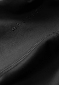 Black athletic fabric with a smooth texture, featuring a subtle embossed brand logo and flat seams, showing a close-up detail.