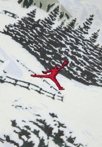 Tkanina s printom koja prikazuje zimski pejzaž s tamnozelenim borovima. Crveni izvezeni Jumpman logo ističe se u prvom planu.