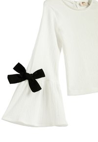 White textured top featuring long bell sleeves, a flared hem, and a black bow accent on the left sleeve.