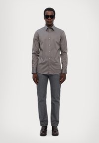 Chemise à bouton gris avec un col classique, associée à un pantalon en denim foncé et des chaussures noires brillantes. Le modèle porte des lunettes de soleil sombres.