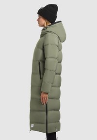 Manteau matelassé vert olive au design long, avec capuche, zips verticaux noirs et bonnet noir côtelé. Texture lisse, silhouette mi-genoux.