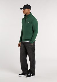 Groene fleece trui met een ritskraag, gecombineerd met donkere jeans en zwarte sneakers. De pet is zwart met geborduurde details.