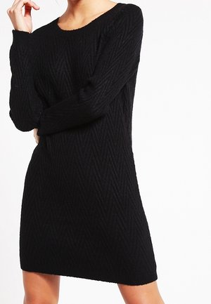 Femme portant une robe noire en tricot à manches longues avec un motif chevron, debout avec un bras croisé et l'autre main près du visage.