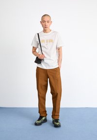 Weißes T-Shirt mit goldfarbenem "AIR"-Logo, braune Hose und schwarze Umhängetasche. Sneaker mit grünem und schwarzem Muster. Einfarbiger Hintergrund.