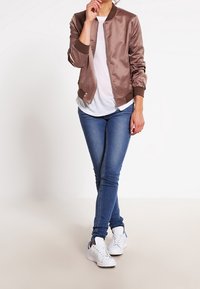 Světle hnědá saténová bomber bunda se zipem, ribrovanými manžetami a bílým tričkem, v kombinaci s modrými skinny džíny a bílými teniskami.