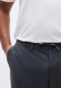 Άνδρας που φοράει σκούρα γκρι παντελόνια με μαύρη ζώνη δεμένη φιόγκο, λευκό πουκάμισο μπουζωμένο και το αριστερό χέρι στην τσέπη, σε απλό φόντο.