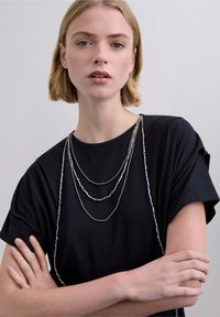 Tres cadenas plateadas de varios niveles con distintos acentos de cuentas caen sobre una camiseta negra de cuello redondo, con mangas cortas y un ajuste relajado.