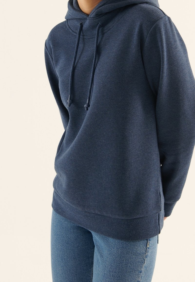 Personne portant un sweat à capuche bleu foncé et un jean bleu, debout devant un fond clair uni.