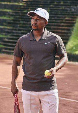 Camisa de polo a rayas marrón con un logo de Nike, combinada con pantalones blancos. Sosteniendo una pelota de tenis amarilla y una raqueta de tenis en una cancha de tierra batida.
