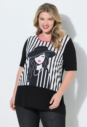 Smilende kvinde med blondt hår iført sorte jeans og en sort-hvid stribet top med grafik af en kvinde i hat og perlekæde.