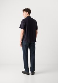 Polo a maniche corte blu navy con taglio dritto, abbinato a jeans in denim blu scuro e scarpe casual nere, visto da dietro.