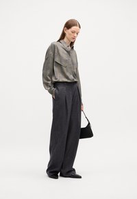 Camicia a scacchi grigia con maniche lunghe e due tasche applicate, abbinata a pantaloni grigi scuri larghi con pieghe e una piccola borsa nera.