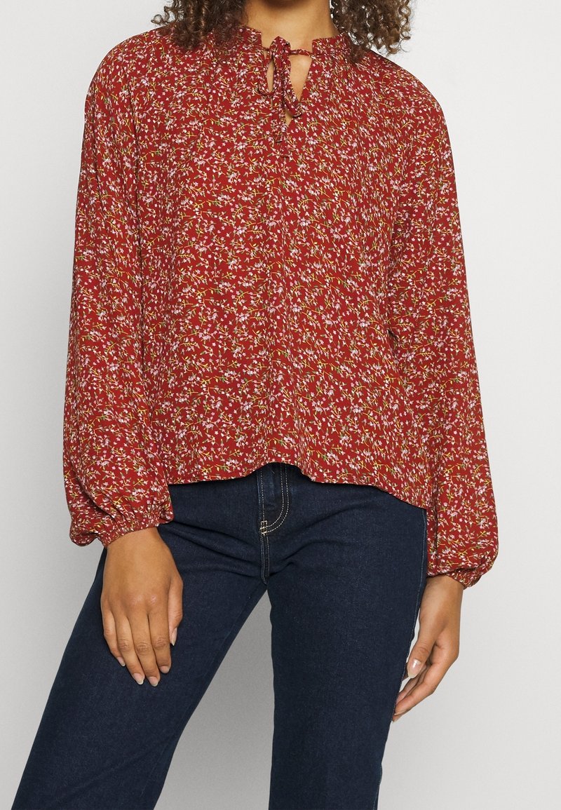 Femme portant un chemisier rouge à manches longues avec un petit motif floral et un col noué, associé à un jean bleu foncé.
