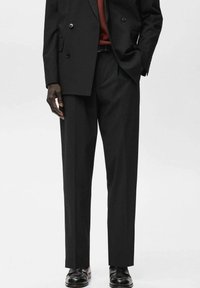 Mężczyzna w czarnym podwójnym żakiecie, dopasowanych spaniach, brązowym pasku i czarnych skórzanych butach, stoi z jedną ręką w kieszeni.